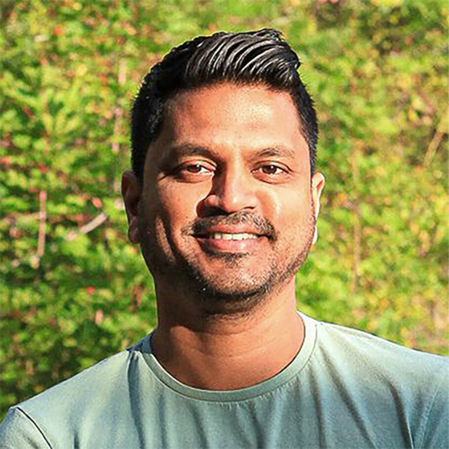 Headshot of Anand Jaggernauth. Anand is a man of West Indian/Caribbean descent, with short black hair and dark eyes. He is smiling, wearing a light green shirt and standing outside with trees in the background.