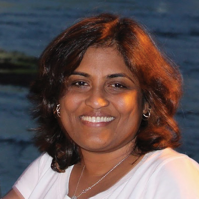 Headshot of Anjana Magapu. Anjana is a woman of South Asian descent with shoulder-length brown hair and brown eyes. She is smiling, wearing a white shirt and sitting in front of the ocean.