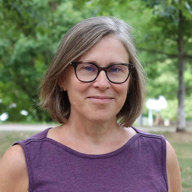 Headshot of Corinne Smirle. Corinne is a White woman with shoulder length light brown and grey hair, blue eyes and wearing glasses. She is standing outside in front of row of trees, and is wearing a purple shirt.