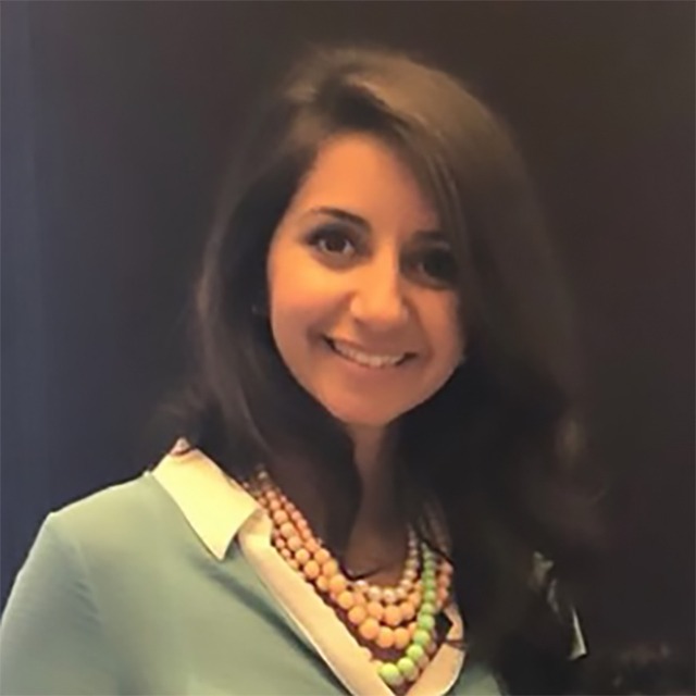 Headshot of Julia Kundacki. Julia is a female person of colour, of Middle Eastern descent. She has dark brown hair and light brown eyes She is wearing a white blouse and light green sweater, and is smiling in front of a dark background.