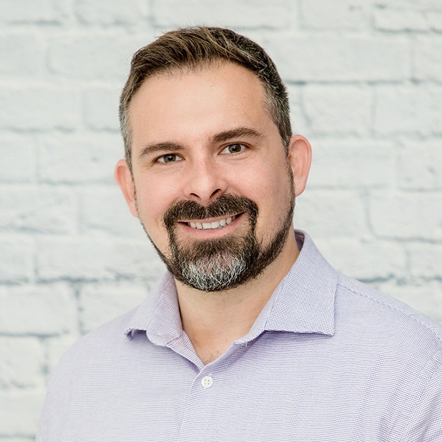 Headshot of Justin Goddard. Justin is a White man with short brown hair, and short facial hair. Justin has flecks of gray in his beard and hair. He is smiling and standing in front of a white-bricked background, wearing a purple button-up shirt.