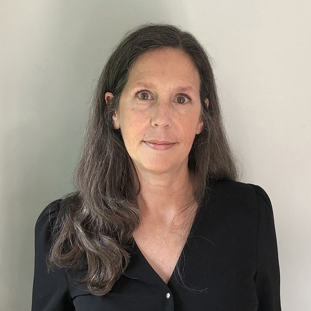 Headshot of Kate Scowen. Kate is a White woman with long brown and grey hair, and brown eyes. She is smiling, wearing a black shirt and standing against a white wall.