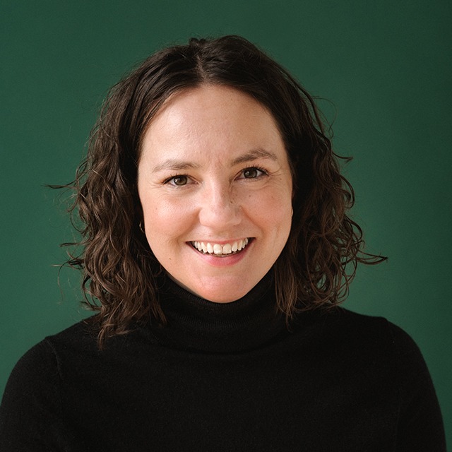 NATASHA2-FULLPROFILE Headshot of Natasha Busic. Natasha is a White woman with curly brown hair, wearing a black sweater, smiling and standing in front of a green background.