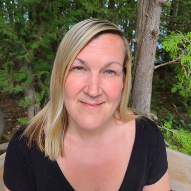 Headshot of Tiina Eldridge. Tiina is a white woman with shoulder length blond hair and green eyes. Tiina is wearing a black v-neck t-shirt and sitting in front of green trees.