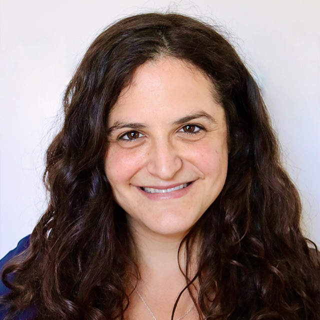 Headshot of Aviva Bellman. Aviva is an Ashkenazi Jewish woman with long brown curly hair, and brown eyes. She is smiling, wearing a blue shirt and standing in front of a white background.