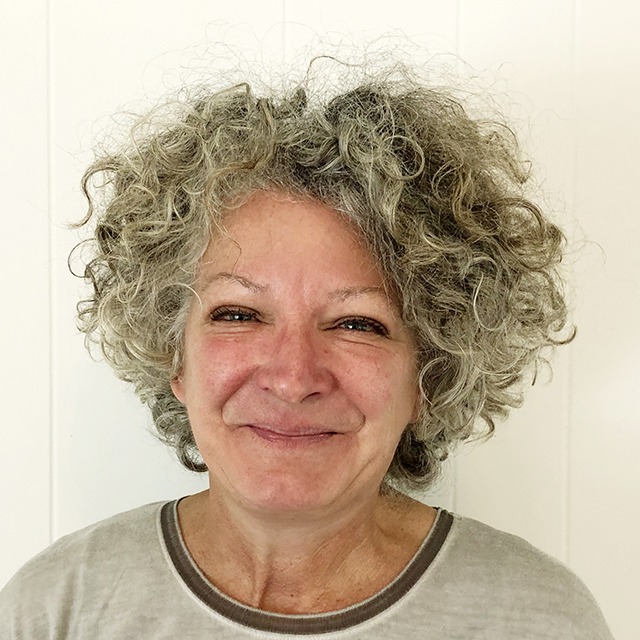 SONYA-THURSBY-FULLPROFILE Headshot of Sonya V. Thursby. Sonya is a white woman with chin-length curly grey and white hair and green-blue eyes, standing in front of a white wall wearing a grey shirt.