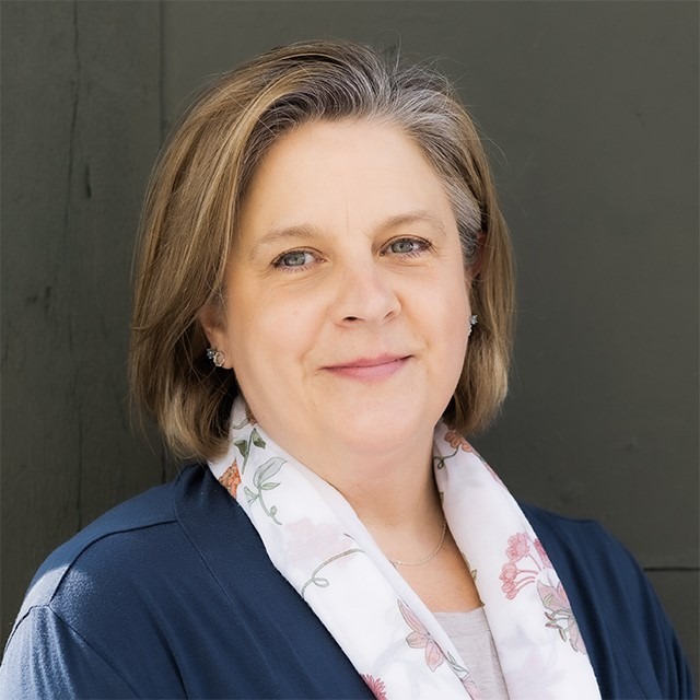 thumbnail_JENNIFER-SERVINIS-NEW-FULLPROFILE Headshot of Jennifer Servinis. Jennifer is a white woman with medium length brown and grey hair and green eyes, standing outside in front of a grey wall and wearing a blue long sleeve sweater with a beige t-shirt underneath and floral pattern scarf around her neck.