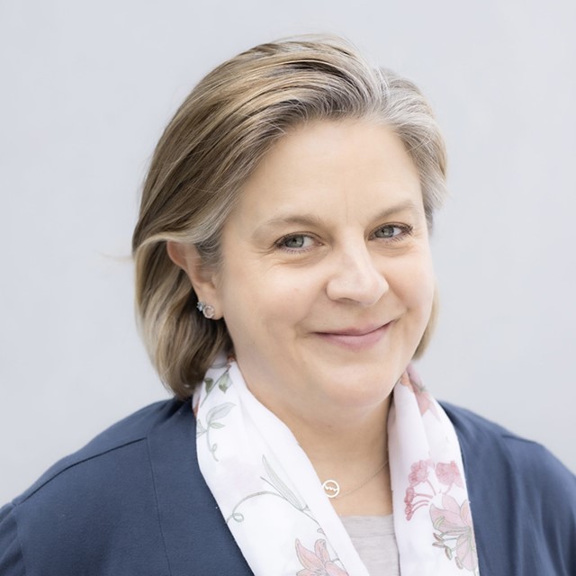 Headshot of Jennifer Servinis. Jennifer is a white woman with medium length brown and grey hair and green eyes, standing outside in front of a light grey wall and wearing a blue long sleeve sweater with a beige t-shirt underneath and floral pattern scarf around her neck.