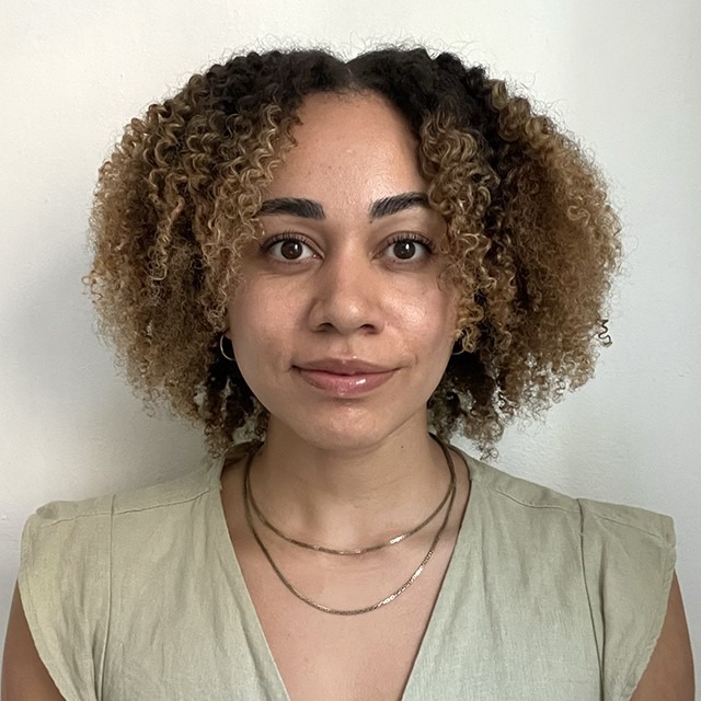 Headshot of Rebekah Otim. Rebekah is a bi-racial Black woman with curly brown and blond hair.  She is standing in front of a white wall and is wearing a green shirt. 