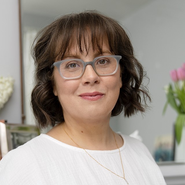 Headshot of Caroline Lock. Caroline is a white woman wearing blue glasses. She has brown wavy shoulder-length hair with bangs. She is wearing a white shirt.