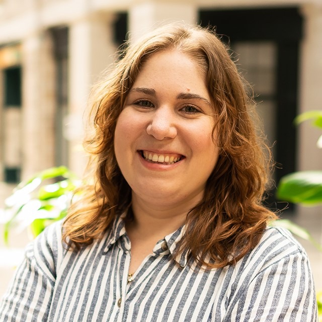 Jessica is a white woman with green eyes and dark blonde hair. She is wearing a blue and white striped collared shirt, smiling warmly while standing in an atrium with a planter box in the background.