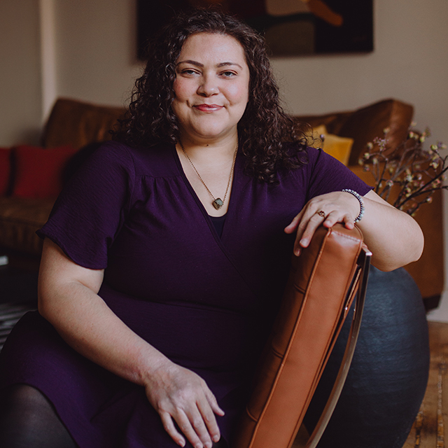 Headshot of Cristina Fuller. Cristina is a white woman with shoulder length curly brown hair and green eyes standing in front of a gray and pink wall wearing a dark green shirt.  