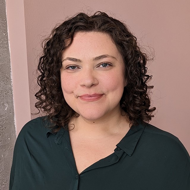 Headshot of Cristina Fuller. Cristina is a white woman with shoulder length curly brown hair and green eyes standing in front of a gray and pink wall wearing a dark green shirt.  