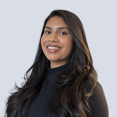 thumbnail_Pirntha-Rajini-FULLPROFILE Headshot of Pirntha Rajini. Pirntha is a Tamil woman with long black hair and brown eyes, smiling with her teeth, standing in front of a light blue wall, and wearing a black turtleneck top.