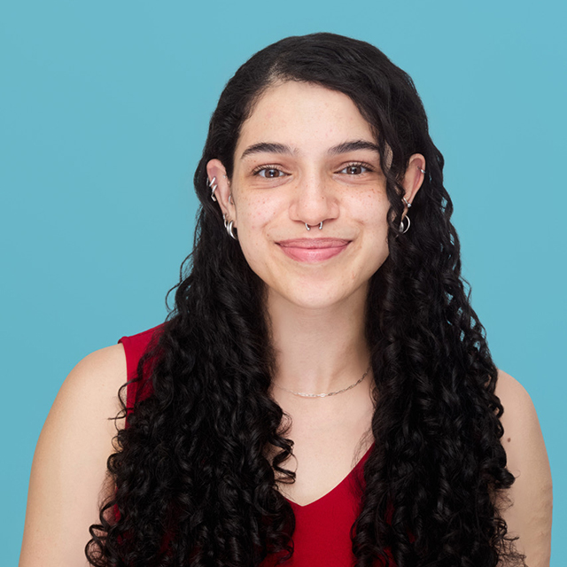 A portrait of Rose -- she is wearing a red shirt, has long dark hair, and is set against a blue background.